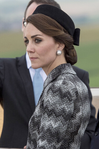 kate middleton wearing a grey outfit with her hair twisted into a low bun with a hairnet accessory and a black hat